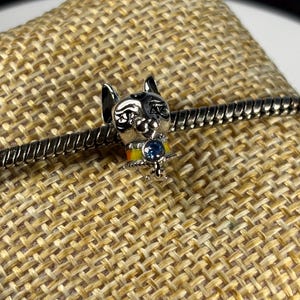 May include: Silver charm featuring a cat with a blue gem on its collar. The cat is wearing a yellow, orange, and red striped collar with a silver charm attached.