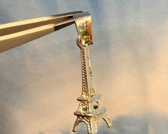 May include: A silver charm in the shape of the Eiffel Tower. The charm is hanging from a pair of tweezers.