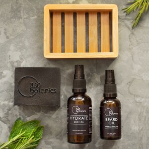 May include: Two amber glass bottles with black pump tops and labels. The bottles are labelled "360 Botanics Hydrate Body Oil" and "360 Botanics Beard Oil". A black bar of soap with the text "360 Botanics" is in the background. A bamboo soap dish is also in the background.