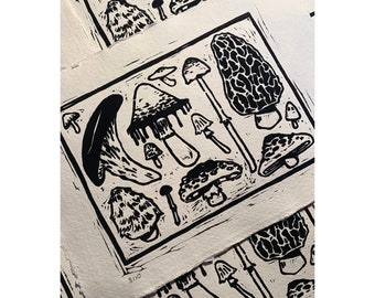 May include: Black and white linocut print featuring various mushroom illustrations. The print showcases a variety of mushroom types, each with unique shapes and textures. The artwork is on textured paper, with the edition number 8/10 visible in the corner.