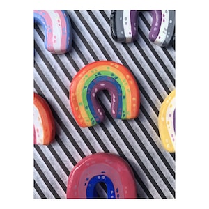 May include: A collection of colorful, hand-painted rainbow-shaped objects. The rainbow arches are painted in various colors, including red, orange, yellow, green, blue, and purple. Some arches have additional colors and patterns. The objects are arranged on a black and white striped background.
