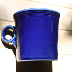 May include: A cobalt blue ceramic mug with a rounded handle. The mug has a cylindrical shape with a slightly flared rim and a light blue accent. The mug is sitting on a wooden surface.
