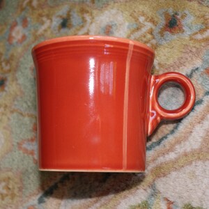 May include: A glossy, orange ceramic mug with a rounded handle. The mug has a cylindrical shape with a slightly flared rim. A vertical, lighter orange stripe runs down the side. The mug is sitting on a patterned rug.