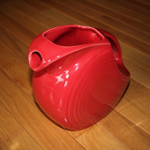 May include: A red ceramic pitcher with a smooth, glossy finish. The pitcher has a wide, round base and a narrow spout.
