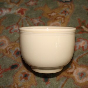 May include: A cream-colored, round ceramic bowl with a slightly flared rim. The bowl has a smooth, glossy finish and sits on a small, circular base. The bowl is likely used for serving food or beverages.