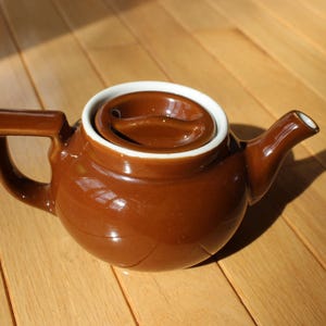 May include: A brown ceramic teapot with a matching lid and a white rim. The teapot has a curved handle and a spout. The teapot is sitting on a wooden surface.