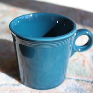 May include: A teal ceramic mug with a rounded handle. The mug has a smooth, glossy finish and a simple, classic design. The mug is sitting on a patterned surface.