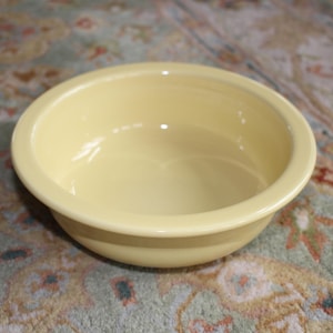 May include: A pale yellow ceramic bowl with a rounded shape. The bowl has a smooth, glossy finish and a slightly raised rim. The bowl is sitting on a patterned rug.