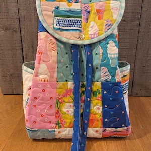 May include: A patchwork backpack with a flap closure and drawstring. The backpack features a variety of colorful fabric patterns, including ice cream cones, flowers, and a typewriter. The straps are blue, and the overall design is whimsical and handmade.
