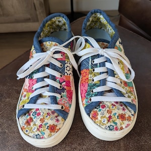 May include: A pair of low-top sneakers featuring a quilted patchwork design with various floral patterns in shades of pink, orange, yellow, and green. The shoes have blue fabric trim, white laces, silver-tone eyelets, and thick white rubber soles.