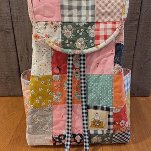 May include: A patchwork backpack with a flap closure and two side pockets. The bag features a variety of fabric squares in floral, gingham, and solid patterns, in shades of pink, orange, green, and blue. A small patch with a chicken illustration is sewn on the front.