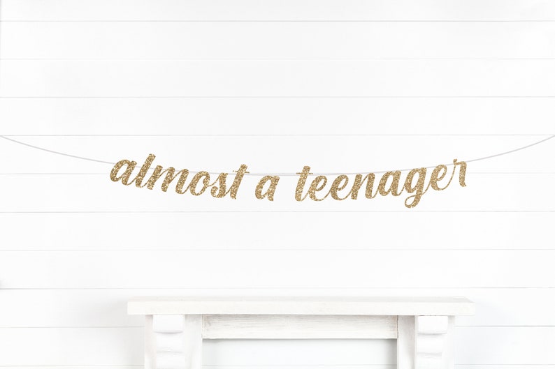May include: Gold glitter banner that reads "almost a teenager" strung across a white background. The banner is a decorative element, likely for a birthday celebration or milestone event. The text is in a cursive font.