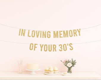 May include: A gold glitter banner that reads "IN LOVING MEMORY OF YOUR 30'S" hangs above a table with a cake, cupcakes, and flowers. The banner is strung across a light pink wall, creating a celebratory atmosphere.