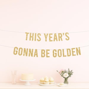 May include: A gold glitter banner that reads "THIS YEAR'S GONNA BE GOLDEN" hangs above a white table with a cake, cupcakes, and flowers. The banner is strung across a light pink wall, creating a festive atmosphere.