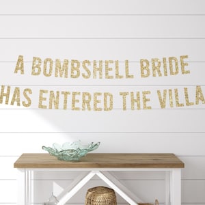 May include: A gold glitter banner with the text "A BOMBSHELL BRIDE HAS ENTERED THE VILLA" hangs on a white shiplap wall. Below, a wooden table holds a decorative glass bowl. A woven basket sits on the shelf below.