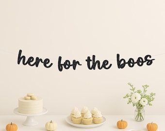 May include: A festive Halloween party scene with a black glitter banner that reads "here for the boos." The table is decorated with a cake, cupcakes, pumpkins, and flowers, creating a celebratory atmosphere.