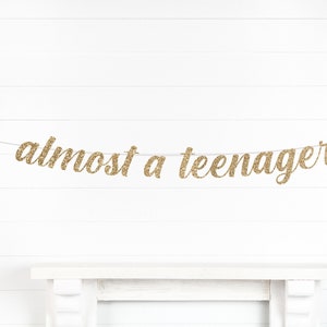 May include: Gold glitter banner that reads "almost a teenager" strung across a white background. The banner is a decorative element, likely for a birthday celebration or milestone event. The text is in a cursive font.