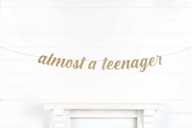 May include: Gold glitter banner that reads "almost a teenager" strung across a white background. The banner is a decorative element, likely for a birthday celebration or milestone event. The text is in a cursive font.