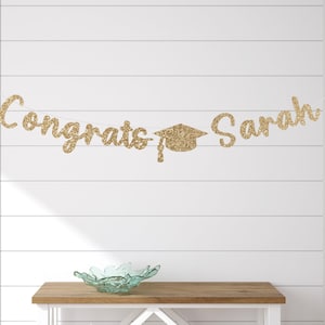 May include: A gold glitter banner that reads "Congrats Sarah" with a graduation cap in the center. The banner is strung across a white wall. A wooden table and decorative bowl are in the foreground.