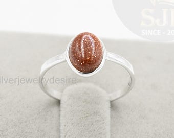 May include: A silver ring featuring an oval, speckled, brown gemstone. The ring has a simple band and a bezel setting that holds the stone. The ring is displayed on a gray surface. The text "Silverjewelrydesire" is visible.