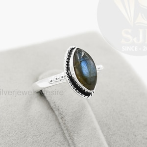 May include: A silver ring featuring an oval-shaped, iridescent gemstone. The gemstone is set in a black bezel with a decorative silver band. The ring is displayed on a gray surface.