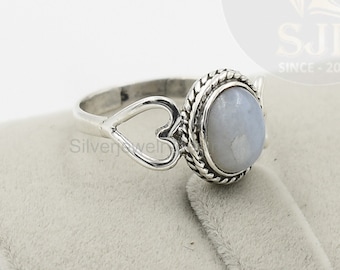 May include: A silver ring with a heart-shaped design and a white oval gemstone. The ring is set on a gray velvet background.