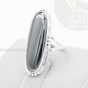 May include: A silver ring featuring a large, elongated, oval-shaped, dark gray stone. The stone is set in a silver band with decorative detailing around the stone and on the ring's shoulders. The ring is displayed on a light gray surface.