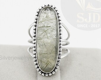 Könnte beinhalten: Ein silberner Ring mit einem länglichen, ovalen Edelstein in hellem Grün mit feinen Einschlüssen. Der Ring hat einen detaillierten, perlenbesetzten Rand und ein geteiltes Schaftdesign. Der Ring wird auf einer grauen Oberfläche präsentiert.