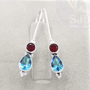 Pode incluir: Brincos de prata com uma pedra preciosa vermelha circular no topo e uma pedra preciosa azul em forma de gota por baixo. Os brincos são exibidos numa superfície cinzenta.