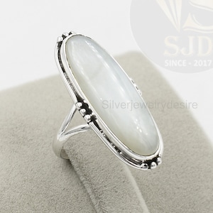 Può includere: Un anello d'argento con una pietra ovale di madreperla bianca incastonata in una ghiera con perline. L'anello è su un espositore di velluto bianco.
