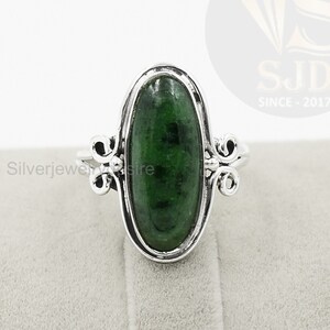 May include: A silver ring with a green oval gemstone set in a bezel setting. The ring has a delicate design with two small swirls on each side of the stone.