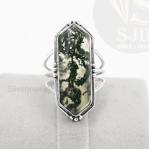 May include: A silver ring featuring a green moss agate stone set in a hexagonal bezel. The ring has a delicate band with a textured finish.