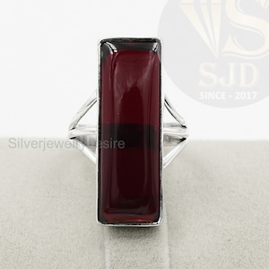 Peut inclure: Une bague en argent avec une pierre précieuse rectangulaire rouge. La bague a un design simple avec deux bandes qui se croisent.