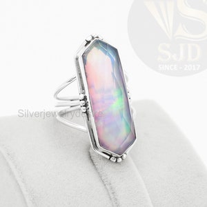 Peut inclure: Une bague en argent avec une grande pierre rectangulaire irisée. La pierre précieuse présente un jeu de couleurs rose, violet et vert. La bague est posée sur un présentoir en velours blanc.