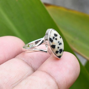 May include: A silver ring with a white and black dalmatian jasper stone. The ring has a simple band with a split design.