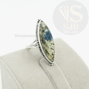 May include: A silver ring with a blue and white oval-shaped stone set in a silver bezel. The ring has a braided band and is on a white velvet display.