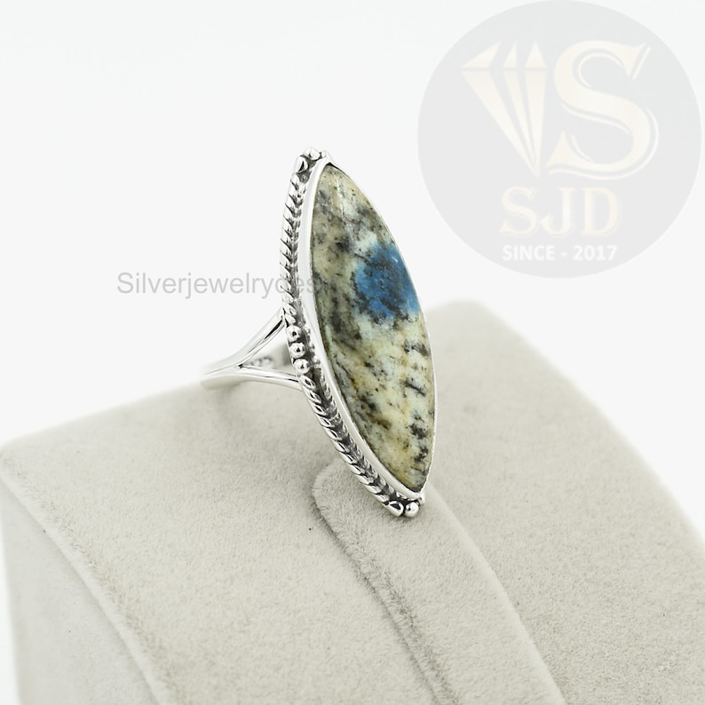 May include: A silver ring with a blue and white oval-shaped stone set in a silver bezel. The ring has a braided band and is on a white velvet display.
