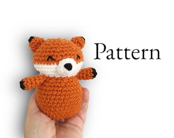 May include: A crocheted orange fox with white accents and black eyes. The fox is holding its paws up and has a happy expression. The word "Pattern" is written in black text on a white background.