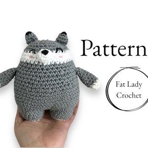 Op de afbeelding: Een grijs gehaakt amigurumi-speelgoed in de vorm van een wolf met witte en roze details. Het speelgoed wordt in de hand van een persoon gehouden. De tekst "Pattern" en "Fat Lady Crochet" is zichtbaar in de afbeelding.