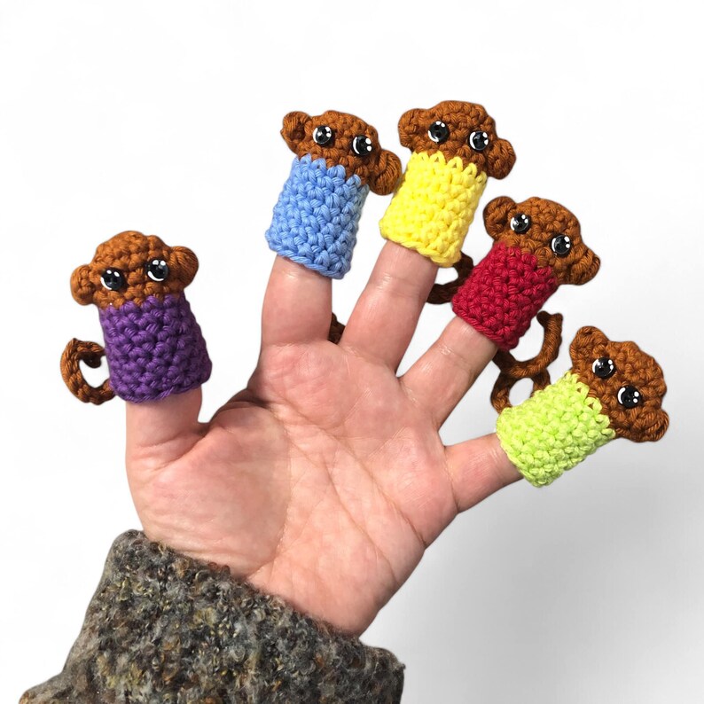 May include: Five finger puppets shaped like monkeys, each wearing a different colored shirt. The monkeys are brown with black eyes and are wearing purple, blue, yellow, red, and green shirts.