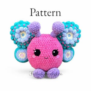 May include: A crocheted butterfly plush toy with pink, purple, and teal colors. The butterfly has large, colorful wings and a smiling face with black eyes. The toy is sitting on a white background. Pattern.