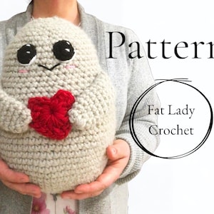 May include: A crocheted plush toy in the shape of a ghost holding a red heart. The ghost has black eyes and a small smile. The toy is being held by a person's hands. The text "Pattern" and "Fat Lady Crochet" is visible in the image.