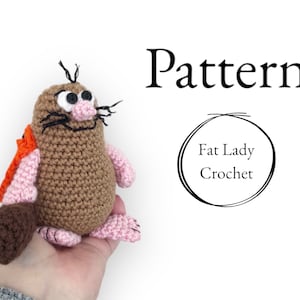 May include: Crocheted mole amigurumi toy with brown fur, pink feet, and a brown tail. The mole has black eyes and a pink nose. The toy is being held in a hand. The text "Pattern" and "Fat Lady Crochet" is visible in the background.