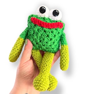 Op de afbeelding: Een handgemaakte gehaakte kikkerpop met grote witte ogen en zwarte pupillen. De kikker heeft een groen lichaam, gele armen en benen en een rode mond. De pop is gemaakt van garen en heeft een getextureerd uiterlijk.