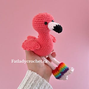 May include: A handmade pink crochet flamingo with a white and black beak, black eyes, and rainbow-colored leg warmers. The flamingo is a whimsical, soft toy, perfect for a child's room or as a decorative item. The text "Fatladycrochet.ca" is visible.