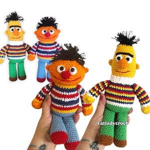 May include: Crocheted Bert and Ernie dolls from Sesame Street. The dolls have yellow and orange skin, black hair, and wear striped tops, green and blue trousers, and brown shoes. The handmade dolls have a whimsical appearance.