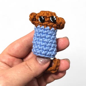 May include: A brown crocheted finger puppet with black eyes and a blue crocheted shirt.