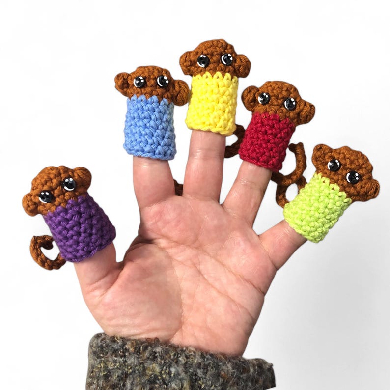 May include: Five finger puppets shaped like monkeys, each wearing a different colored crocheted shirt. The monkeys are brown with black eyes and are wearing purple, blue, yellow, red, and green shirts.