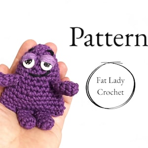 May include: Purple crocheted amigurumi toy with a smiling face and large eyes. The toy is being held in a person's hand. The text "Pattern" and "Fat Lady Crochet" is visible in the image.