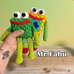 Könnte beinhalten: Zwei handgefertigte Häkelfrosch-Spielzeuge. Einer ist grün mit rotem Mund und gelben Beinen, der andere ist gelb und regenbogenfarben. Beide haben große weiße Augen mit schwarzen Pupillen. Der Text "Mr. Fabu" ist sichtbar.